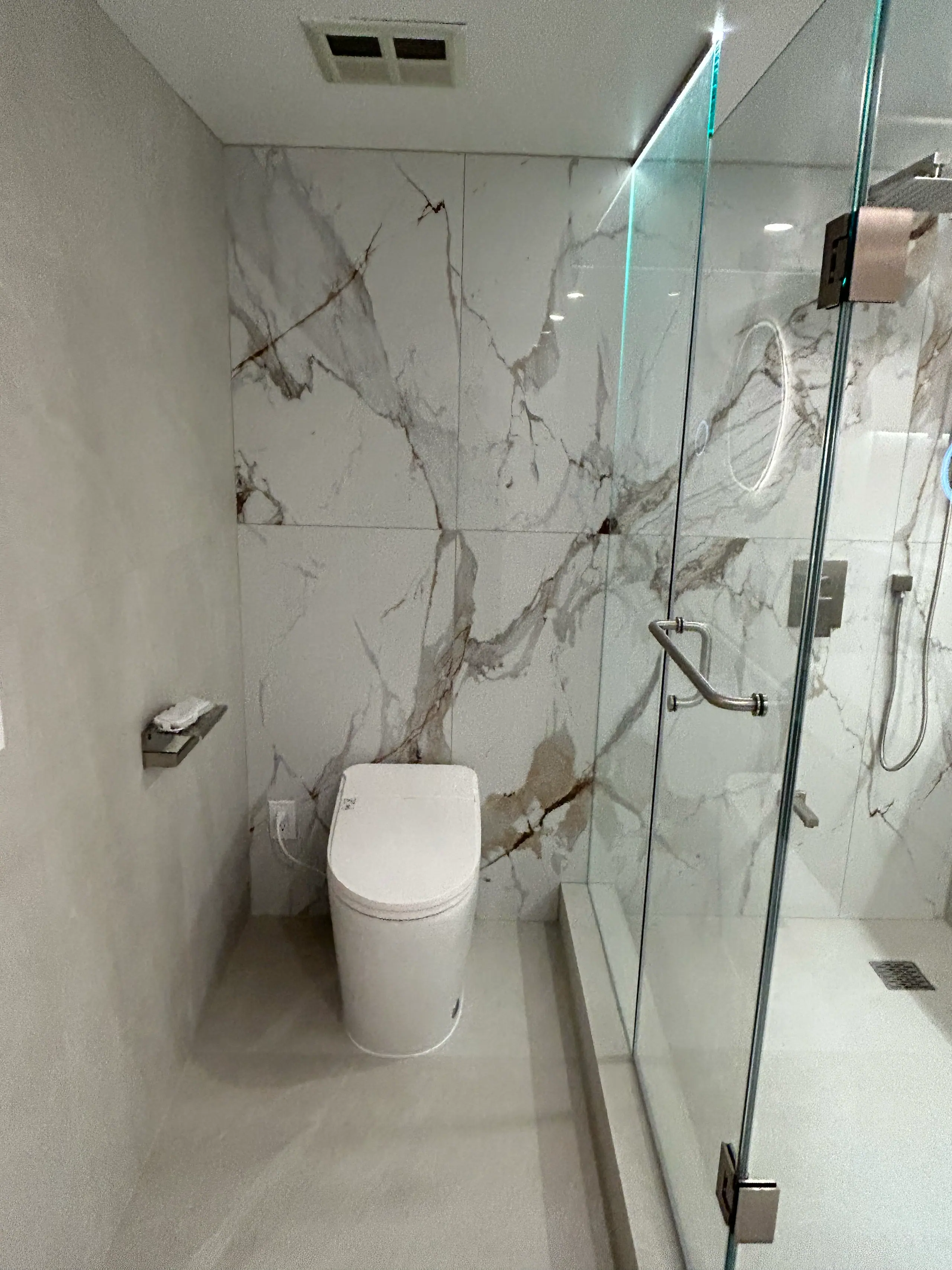 The book-matched Calacatta wall in the finished ensuite running full height behind a concealed-tank toilet, with the glass enclosure and its chrome pull at right and soft grey large-format panels opposite.